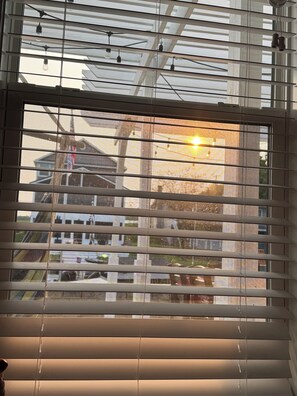 Ein Fenster mit horizontalen Jalousien, die Sonne ist sichtbar, und es bietet Blick auf ein Gebäude.