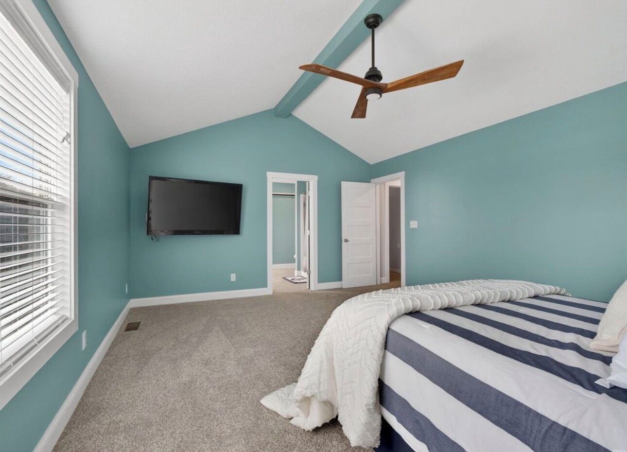 Une chambre à coucher avec un lit, un ventilateur de plafond, un téléviseur fixé au mur et une porte donnant sur une autre pièce.
