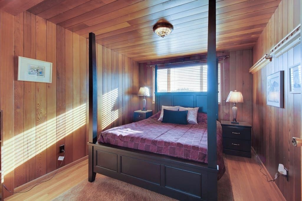 A four-poster bed with a dark frame, a blue headboard, and a purple bedspread. There are two bedside tables with lamps, a window with blinds, and a wall-mounted picture frame.