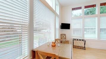 A room with a wooden table and chairs, a flat-screen TV mounted on the wall, and large windows with blinds.
