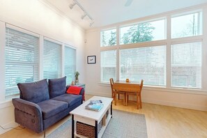 A living room with a sofa, coffee table, and a dining table with chairs. There are large windows with blinds, and a picture frame on the wall.