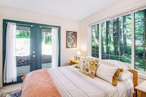 Een slaapkamer met een bed, een nachtkastje, een schilderij en een raam met uitzicht op bomen.