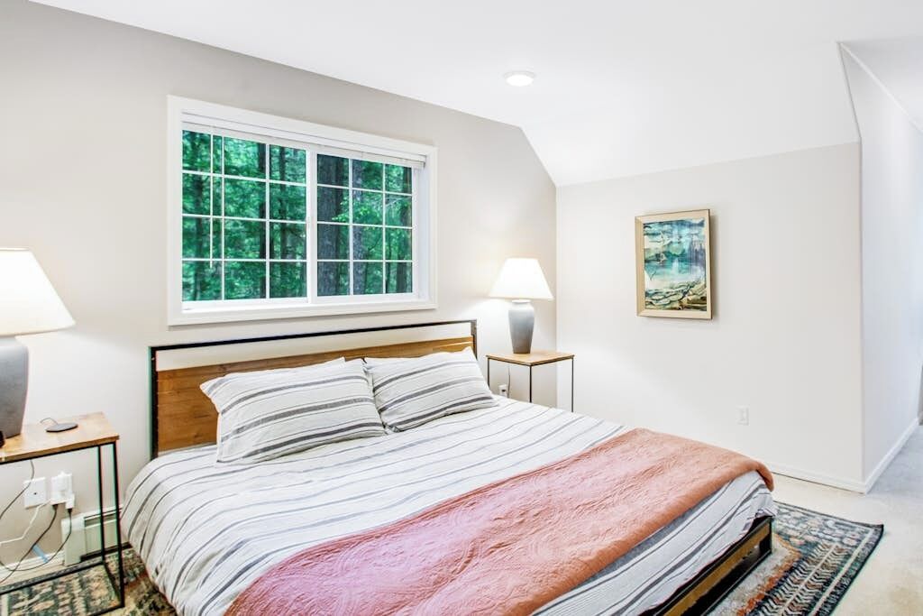 Een slaapkamer met een bed, twee bedlampen, een raam met uitzicht op bomen en een schilderij aan de muur.