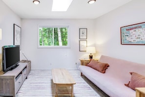 A modern living room with a sofa, a TV, a wooden table, and a window with a view of trees. - Dreamy Retreat in the Woods with Firepit, Fireplace, Free and Large Deck (Eastsound)