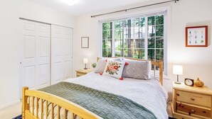 Une chambre à coucher avec un lit en bois, une fenêtre donnant sur des arbres, une table de chevet avec une lampe et un tableau encadré au mur.