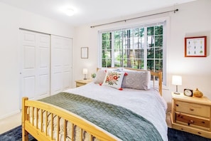 A bedroom with a wooden bed, a window with a view of trees, a bedside table with a lamp, and a framed picture on the wall. - Dreamy Retreat in the Woods with Firepit, Fireplace, Free and Large Deck (Eastsound)