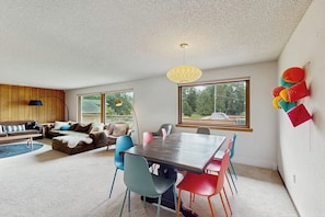 A modern living room with a dining table, colorful chairs, a sofa, and a large window with a view of greenery.
