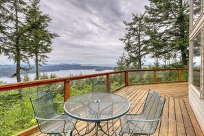 A wooden deck with metal chairs and a table, overlooking a lake and forested mountains.