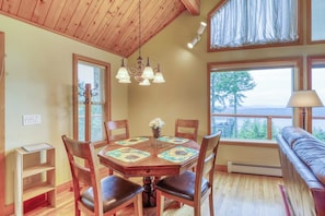 A dining area with a wooden table, chairs, and a chandelier. There is a window with a view of trees and a bench in the background. - Updated Eastsound Home with Stunning Mountain and Ocean Views (Eastsound)