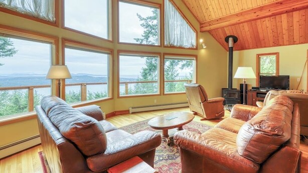 Ein gemütliches Wohnzimmer mit Ledersofas, einem Couchtisch und einem Kamin. Große Fenster bieten Blick ins Freie.