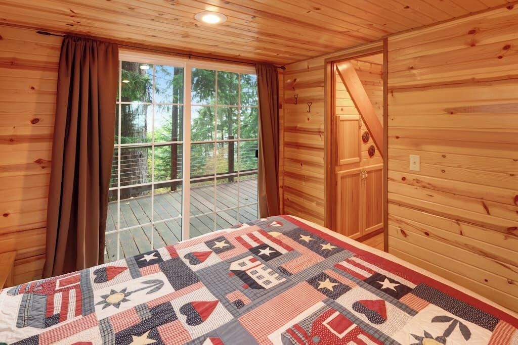Una camera da rifugio in legno con un'ampia finestra, una coperta sul letto e una porta che dà sull'esterno.