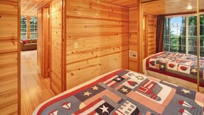A wooden cabin interior with a large bed, a window with a view of trees, and a door leading to another room. - Cozy 1BR Waterfront Cabin with Mountain Views (Eastsound)
