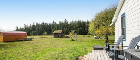 Una terraza de madera con asientos al aire libre, un granero y un cobertizo pequeño en un campo de césped con árboles de fondo.