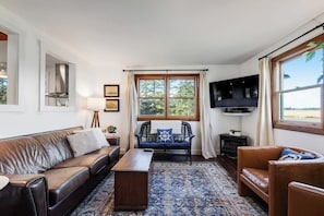 A living room with a sofa, a coffee table, a patterned rug, a window with curtains, and a flat-screen TV.