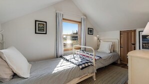 Ein kleines Zimmer mit einem Einzelbett, geneigter Decke, Fenster mit Vorhängen, Nachttisch und einem gerahmten Bild an der Wand.