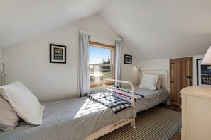 Quarto pequeno com cama de solteiro, teto inclinado, janela com cortinas, mesa de cabeceira e quadro na parede.