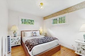 A bedroom with a wooden bed, two bedside tables with lamps, a ceiling light fixture, and two windows with white frames. - Whimsical 1940s Waterview Gem with Sweeping sea views, Wildlife, & Near Town (Langley)