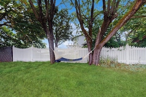 A hammock between trees in a grassy yard with a white picket fence in the background.