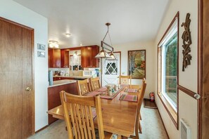 A dining area with a wooden table and chairs, a kitchen in the background, and a window with a view. - Waterfront Cabin with Hot Tub and Mountain Views in Eastsound (Eastsound)