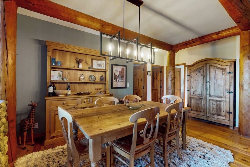 A dining area with a wooden table and chairs, a chandelier, and a rustic wooden cabinet with shelves.