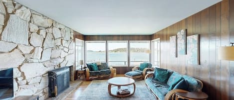 A cozy living room with a stone wall, wooden paneling, and a large window overlooking a body of water.