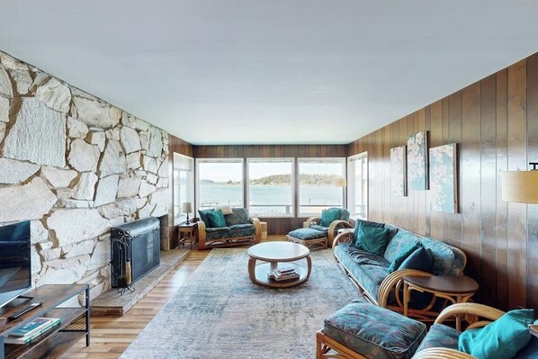 A cozy living room with a stone wall, wooden paneling, and a large window overlooking a body of water.