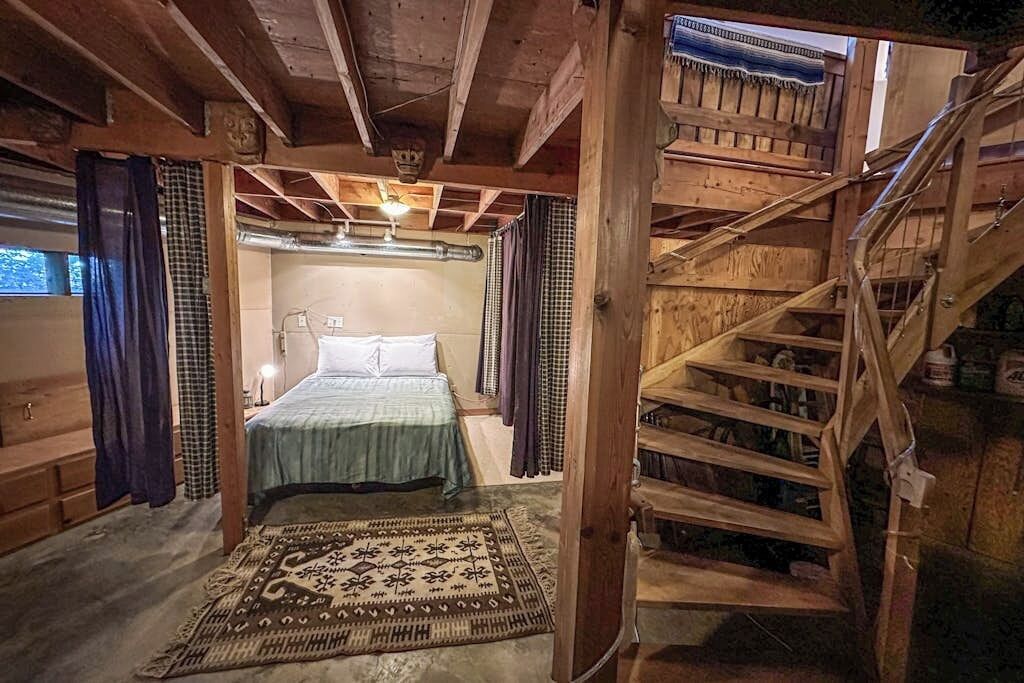 A wooden interior with a bed, a patterned rug, and a staircase.