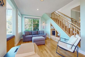 Uma sala de estar com sofá seccional, apoio para os pés e mesa de centro de vidro. Uma escada com corrimãos de madeira leva a um andar superior.