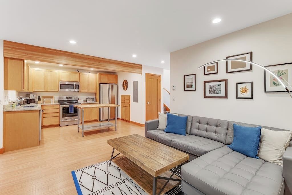 A modern kitchen with wooden cabinets, stainless steel appliances, and a dining area with a grey sofa and blue cushions.