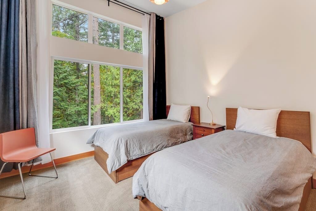 Ein Hotelzimmer mit zwei Betten, einem Schreibtisch und einem Stuhl. Ein großes Fenster bietet Blick auf Bäume.