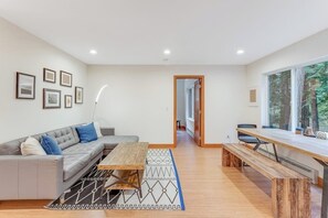 Un saló modern amb un sofà seccional gris, una taula de centre de fusta, i una taula de menjador amb bancs. Hi ha quadres amb marc a la paret i una finestra gran que ofereix vistes a arbres.