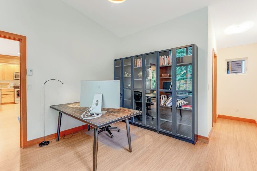 Ein Zimmer mit Holzboden, einem Schreibtisch mit Computer, einem Bücherregal und einer Tür, die in eine Küche führt.