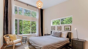 Un dormitorio con una cama grande, una silla, una mesita de noche y un ventanal con vista a árboles.