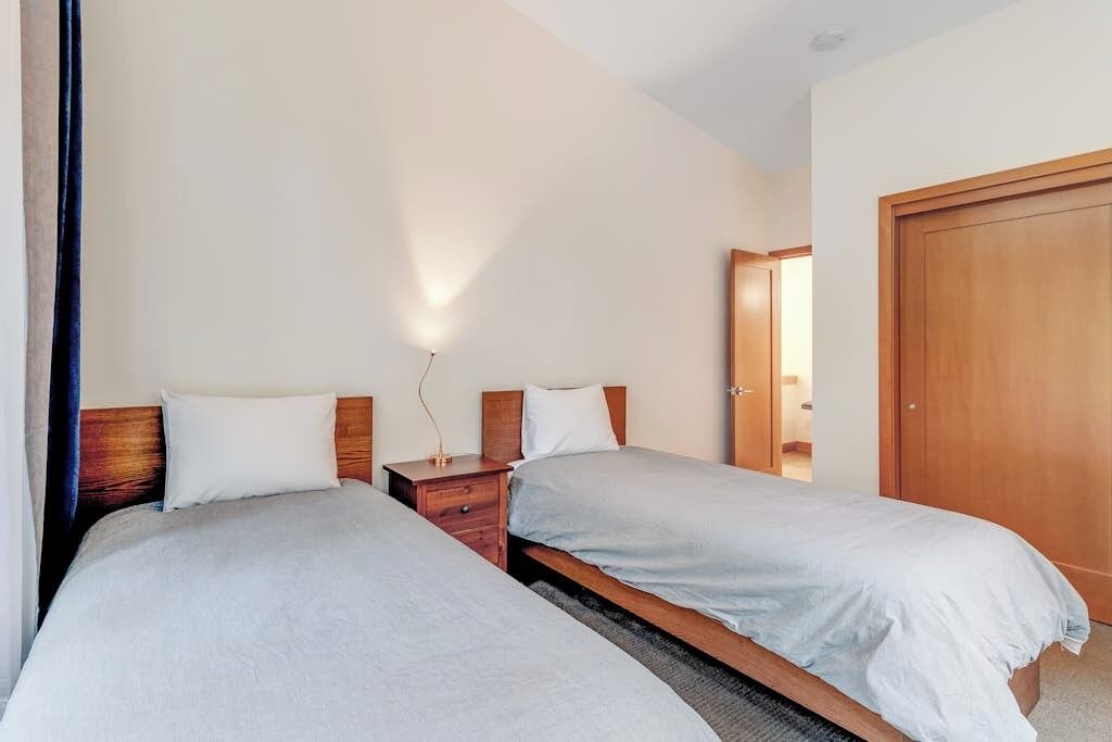 A hotel room with two single beds, wooden headboards, a nightstand with a lamp, and a wooden door.