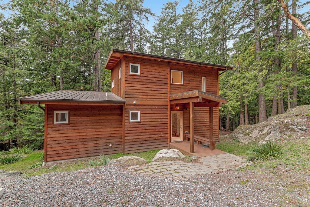 A two-story wooden cabin with a covered porch, surrounded by a forested area.