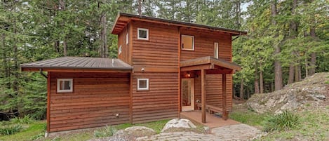 A two-story wooden cabin with a covered porch, surrounded by a forested area.