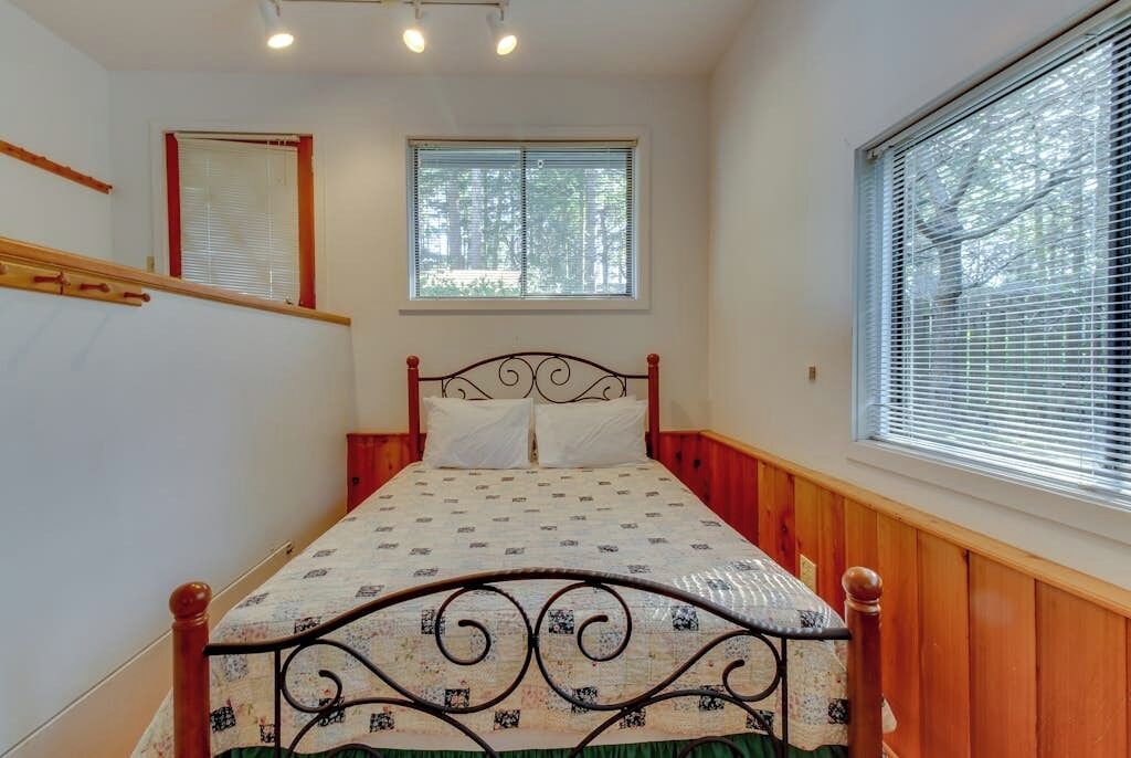 A bedroom with a bed, two windows, and a wooden wall.