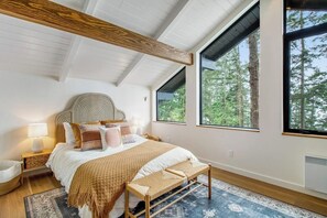 A bedroom with a large bed, a wooden headboard, a bench, a nightstand, and a window with a view of trees. - Stylish Waterfront Oasis with Hot Tub, Fiber Optic Internet, and Entertainer's Deck (Lopez Island)