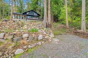 A modern cabin with a stone retaining wall, surrounded by a forested area. - Stylish Waterfront Oasis with Hot Tub, Fiber Optic Internet, and Entertainer's Deck (Lopez Island)
