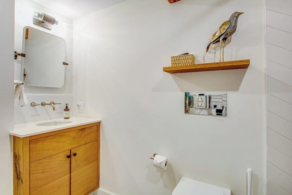 Bany amb un envàs de fusta, un vàter blanc, un mirall, una tassa i una prestatgeria amb un ocell decoratiu.