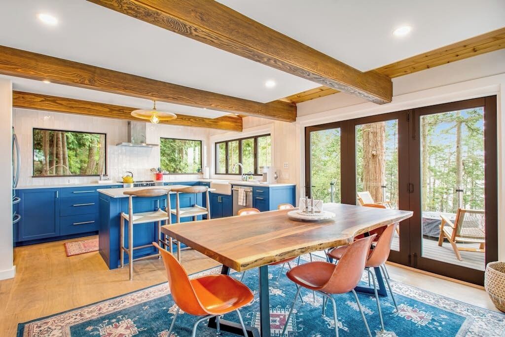 A modern kitchen with blue cabinets, a wooden dining table, and a large window overlooking greenery.