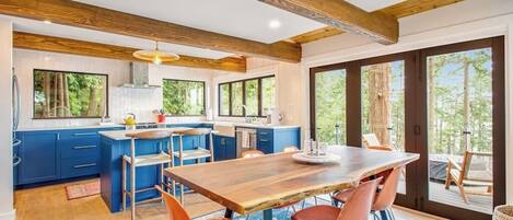 A modern kitchen with blue cabinets, a wooden dining table, and a large window overlooking greenery.