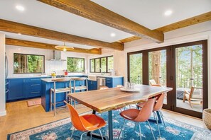 A modern kitchen with blue cabinets, a wooden dining table, and a large window overlooking greenery. - Stylish Waterfront Oasis with Hot Tub, Fiber Optic Internet, and Entertainer's Deck (Lopez Island)