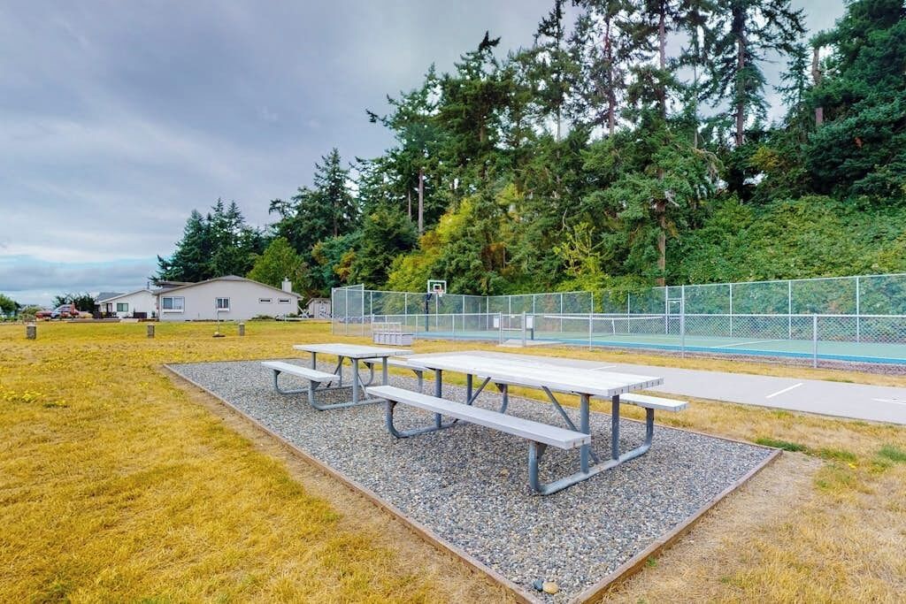 An outdoor area with picnic tables, a basketball court, and a fenced area with trees.
