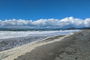 Une plage avec des vagues, des galets et des montagnes lointaines, sous un ciel nuageux.