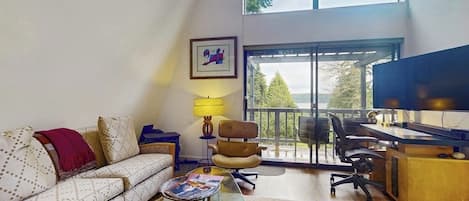 A living room with a sofa, a coffee table, a desk with a computer, and a chair. There is a window with a view of trees and a balcony.