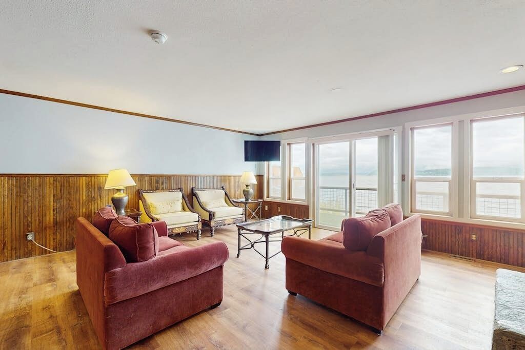 A spacious living room with a wooden floor, two red sofas, a glass coffee table, a white armchair, and a television mounted on the wall.