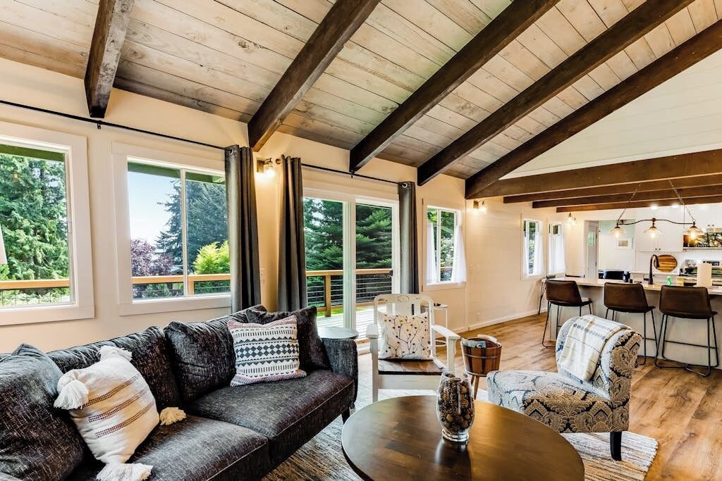 Ein großzügiger Wohnbereich mit einem dunkelgrauen Sofa, einem runden Couchtisch und einem Esstisch mit Stühlen. Große Fenster sorgen für Tageslicht und Blick auf die grüne Umgebung.