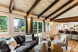 A spacious living area with a dark-colored sofa, a round coffee table, and a dining table with chairs. Large windows provide natural light and views of greenery outside. - Fantastic Waterfront Retreat in Eastsound with Expansive Deck and Stylish Interior - Dog Friendly (Eastsound)