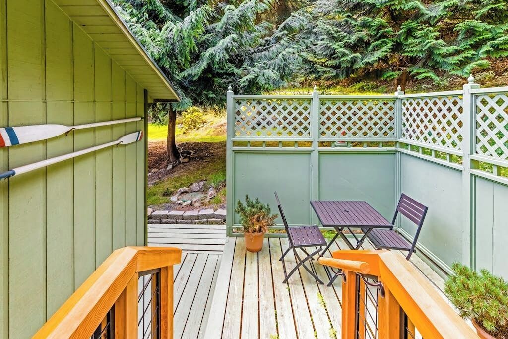 En träveranda med ett bord och stolar, omgiven av ett grönt staket och tät vegetation.